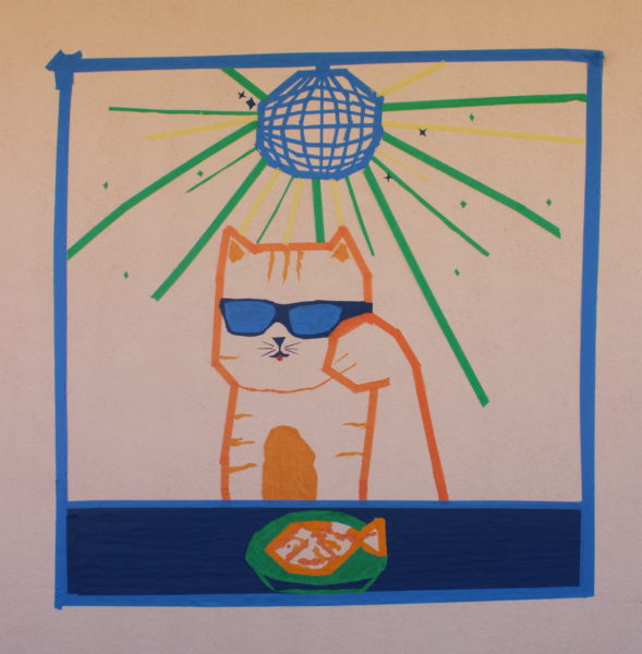 An Innovate mural on a blank wall. The entire thing is outlined by a blue rectangle. At the bottom is a horizontal stripe, outline in light blue and colored in with dark blue, with an oval of green in the center. On the green oval is an orange and white fish. Above this stripe, in the center, is a cat outlined with orange, having multiple orange stripes, and a filled out orange belly. The cats left paw is almost eye-level. The cats pink tongue is sticking out. The cat is wearing sunglasses, the rims in dark blue and the glass in light blue. Above the cat is a disco ball done in light blue, with green and yellow tape outstretching from it, none overlapping with the cat. There are multiple black sparkles, along with green ones, both done in tape.