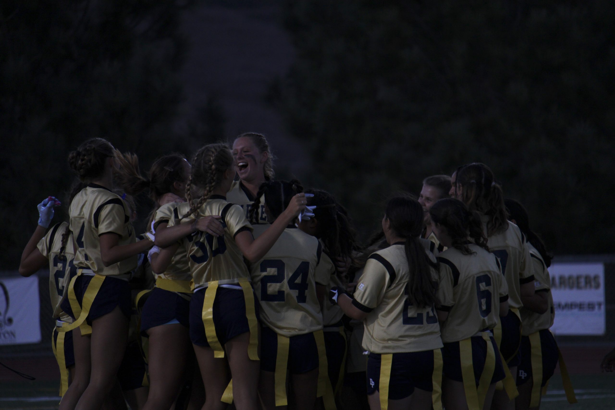 Photo of DPs flag football team at night. The team is in a circle, with most having their arms around each other. All are smiling.